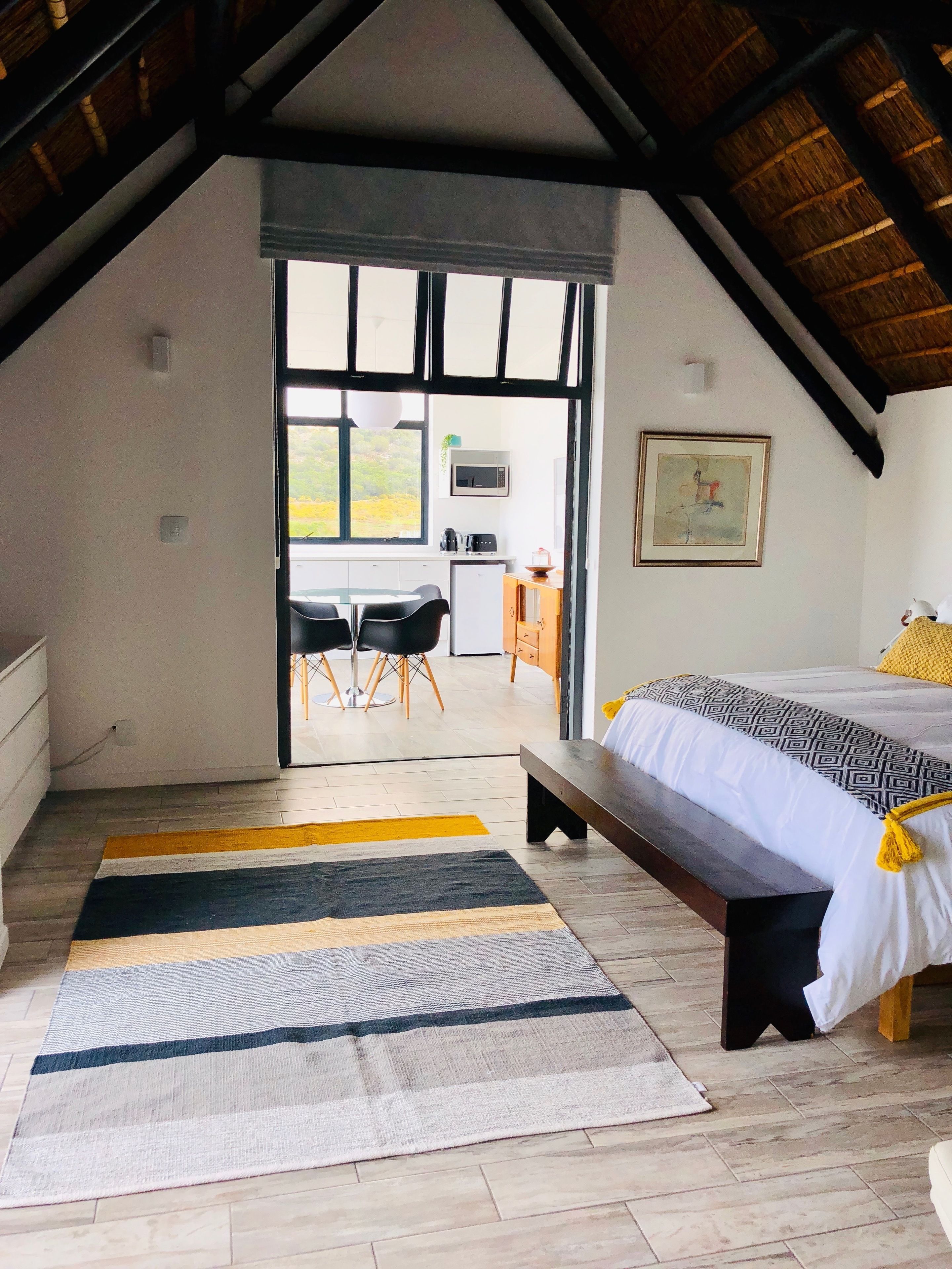 A bedroom with a large bed, a bench, a dining table with chairs, a kitchen area, and a view through a glass door.