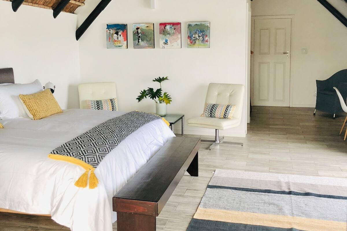 A bedroom with a large bed, a bench, a chair, a small table, and a potted plant.