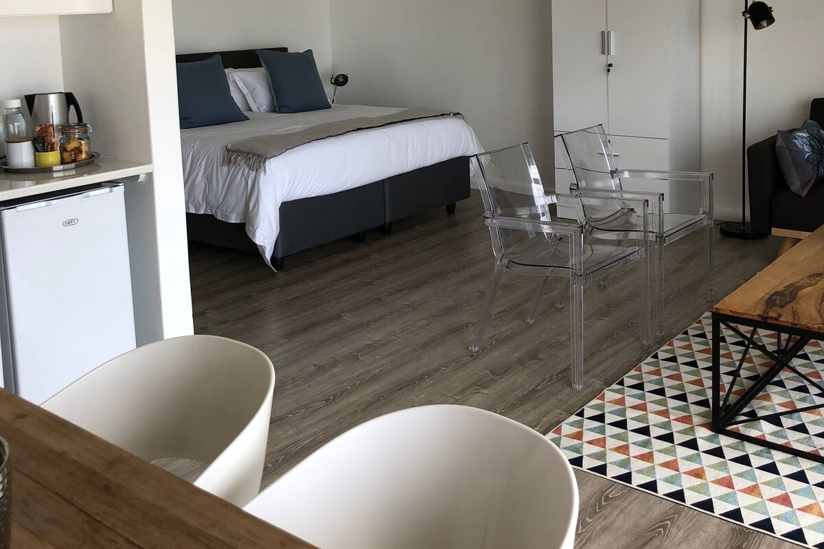 A modern bedroom with a bed, bedside table, and a wardrobe. There is a desk with a chair and a lamp. The room has a wooden floor and a patterned rug.