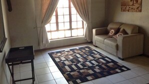 A living room with a sofa, a patterned rug, a window with curtains, and a painting on the wall. - Matalatala (Cullinan)