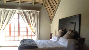 A bed with white linens, two pillows, a bedside table with a vase, and a window with curtains. - Matalatala (Cullinan)