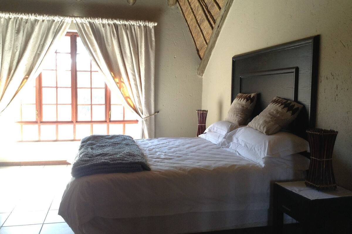 Ein Bett mit weißen Leinentüchern, zwei Kissen, ein Nachttisch mit einer Vase und ein Fenster mit Vorhängen.