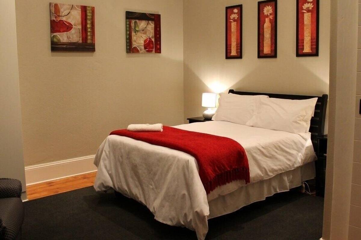 Een netjes opgemaakt bed met wit en rood beddengoed, een zwart hoofdbord en een rode plaid. Aan de muur hangen drie kunstwerken in lijsten, en er staat een klein nachtkastje met een lamp.