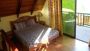 Un dormitorio con techo de madera, una cama con colcha estampada, una mesita de noche con lámpara y vistas a la vegetación a través de un ventanal corredero.