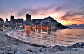 A coastal cityscape with a beach at sunset, featuring modern buildings and a calm sea. - Ocana Apartments (Finestrat)