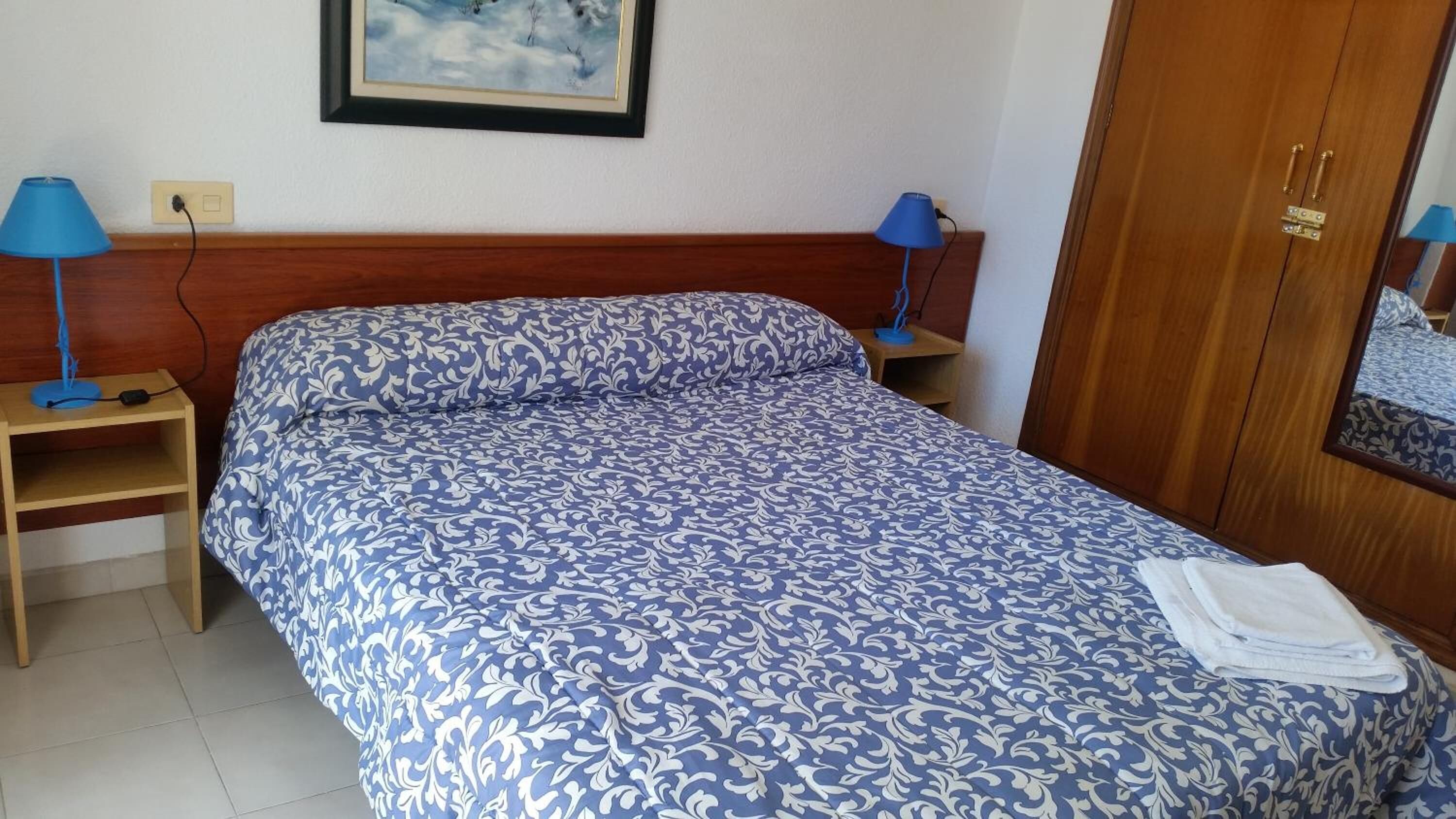 Een slaapkamer met een bed, twee nachtkastjes met lampen, een houten garderobe en een lijst met een afbeelding aan de muur.