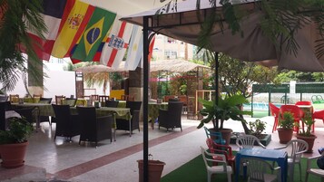 An outdoor dining area with tables and chairs, surrounded by potted plants and flags.