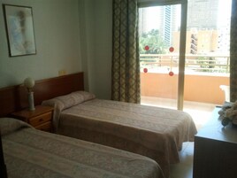 Habitación de hotel con dos camas, una mesita de noche, una lámpara y vistas a un balcón con decoraciones esféricas rojas.