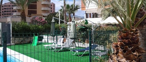 A swimming pool with a green artificial turf area, lounge chairs, and palm trees.