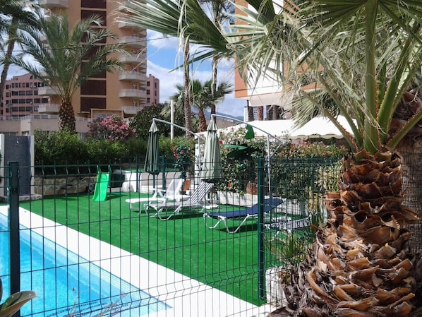 A swimming pool with a green artificial turf area, lounge chairs, and palm trees.