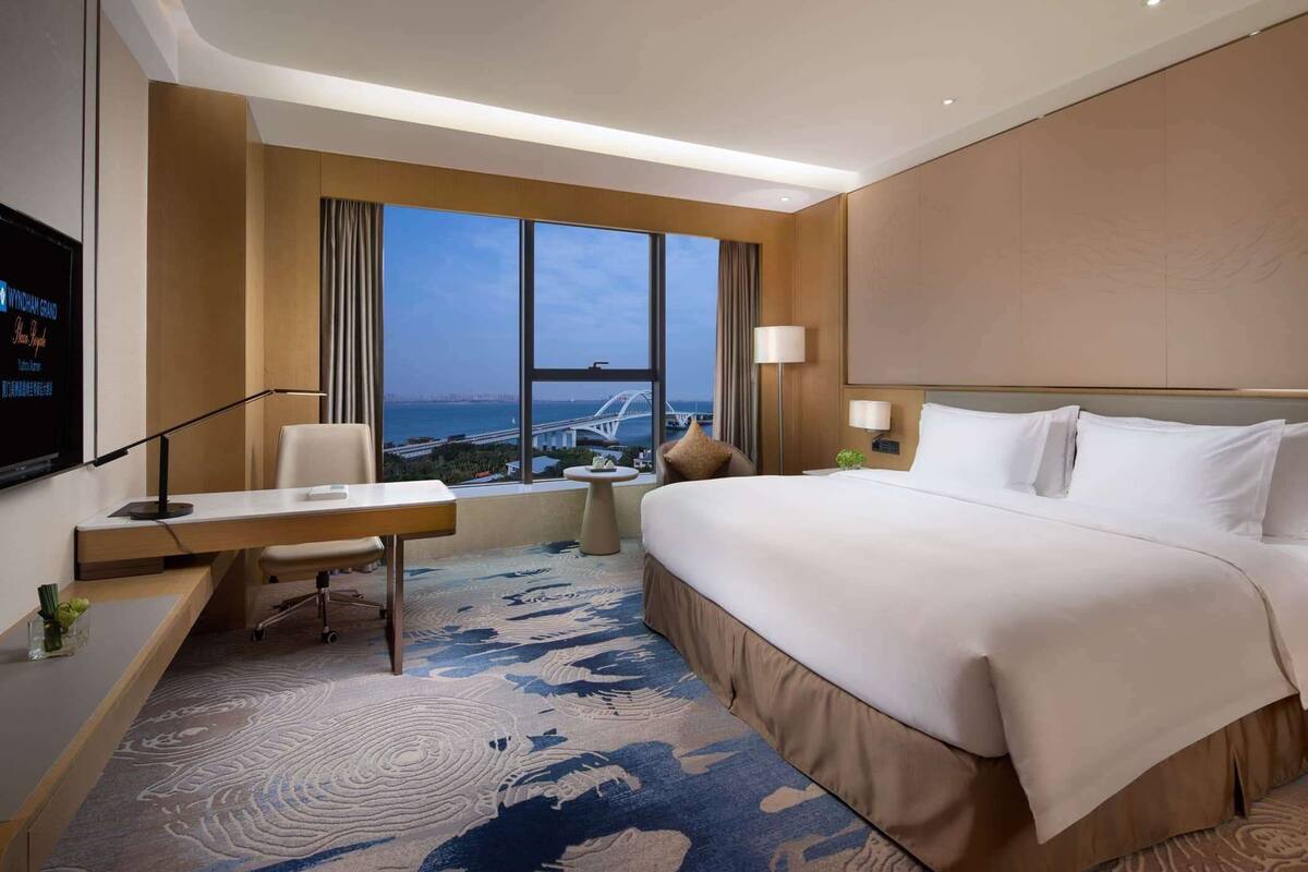 A hotel room with a large bed, a desk with a chair, a TV, and a view of a bridge and cityscape.