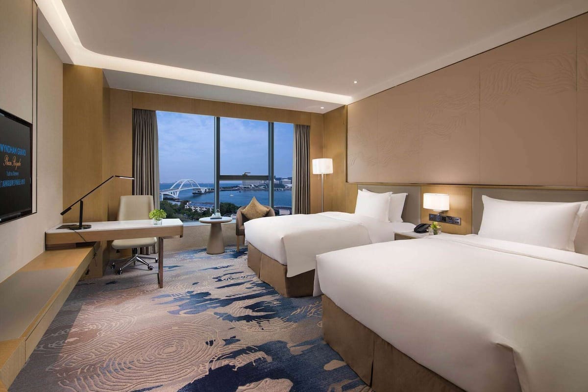 A hotel room with two beds, a desk, a chair, a small table, and a large window offering a view of a bridge and cityscape.