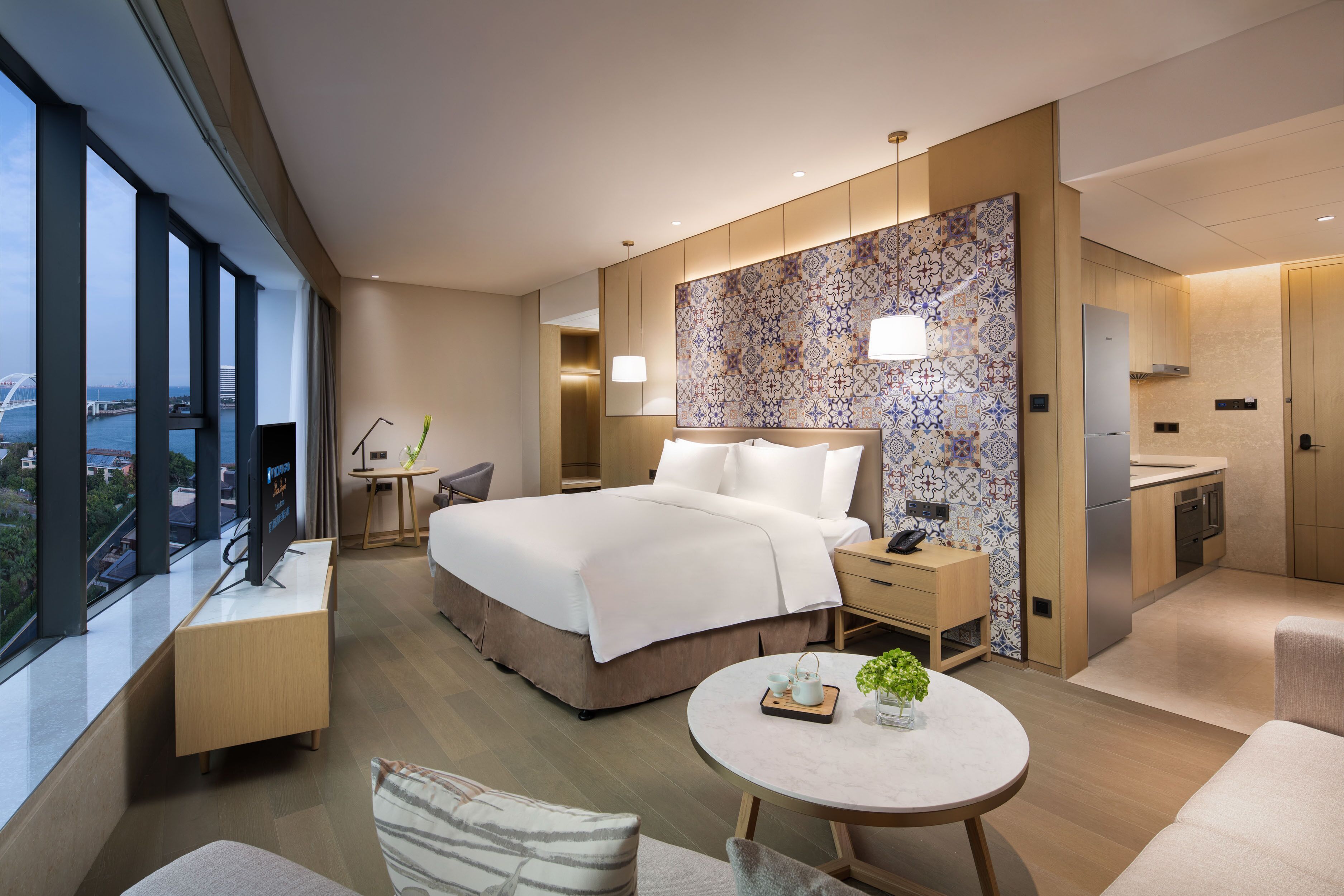 Habitación de hotel moderna con una cama grande, un escritorio, una silla, una pequeña mesa con una planta y vistas a un puente y al agua.