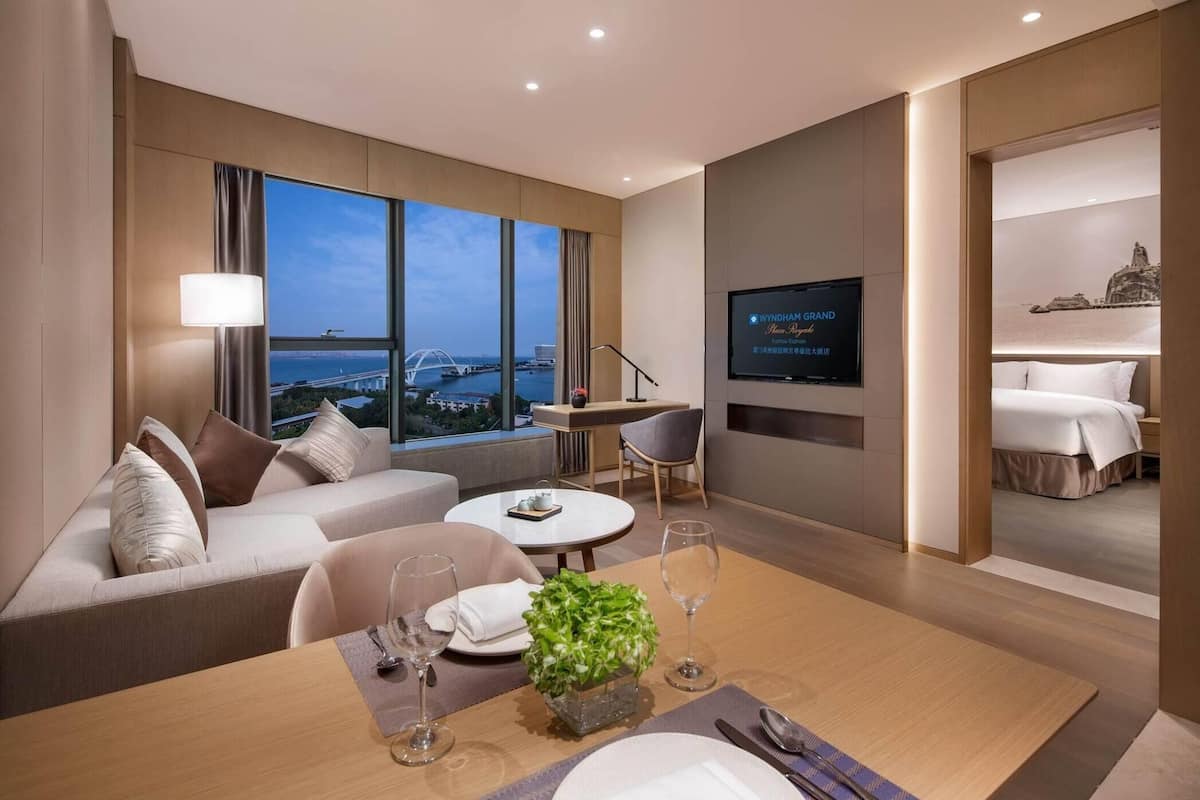 A modern hotel room with a sofa, dining table, TV, and a view of a bridge.