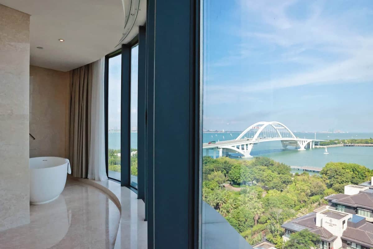 A modern bathroom with a large bathtub, a curved window, and a view of a bridge and greenery.