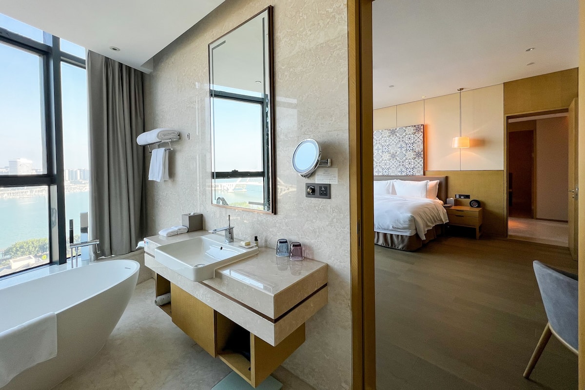 A modern hotel bathroom with a large sink, mirror, and a view of the city.