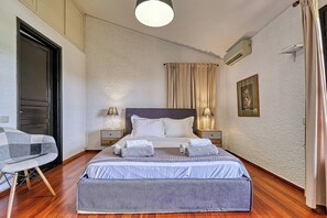 A bedroom with a bed, two bedside tables, a nightstand, a chair, a hanging light fixture, a wall-mounted air conditioner, and a framed picture. - Orange Grove Villa (Nafplio)