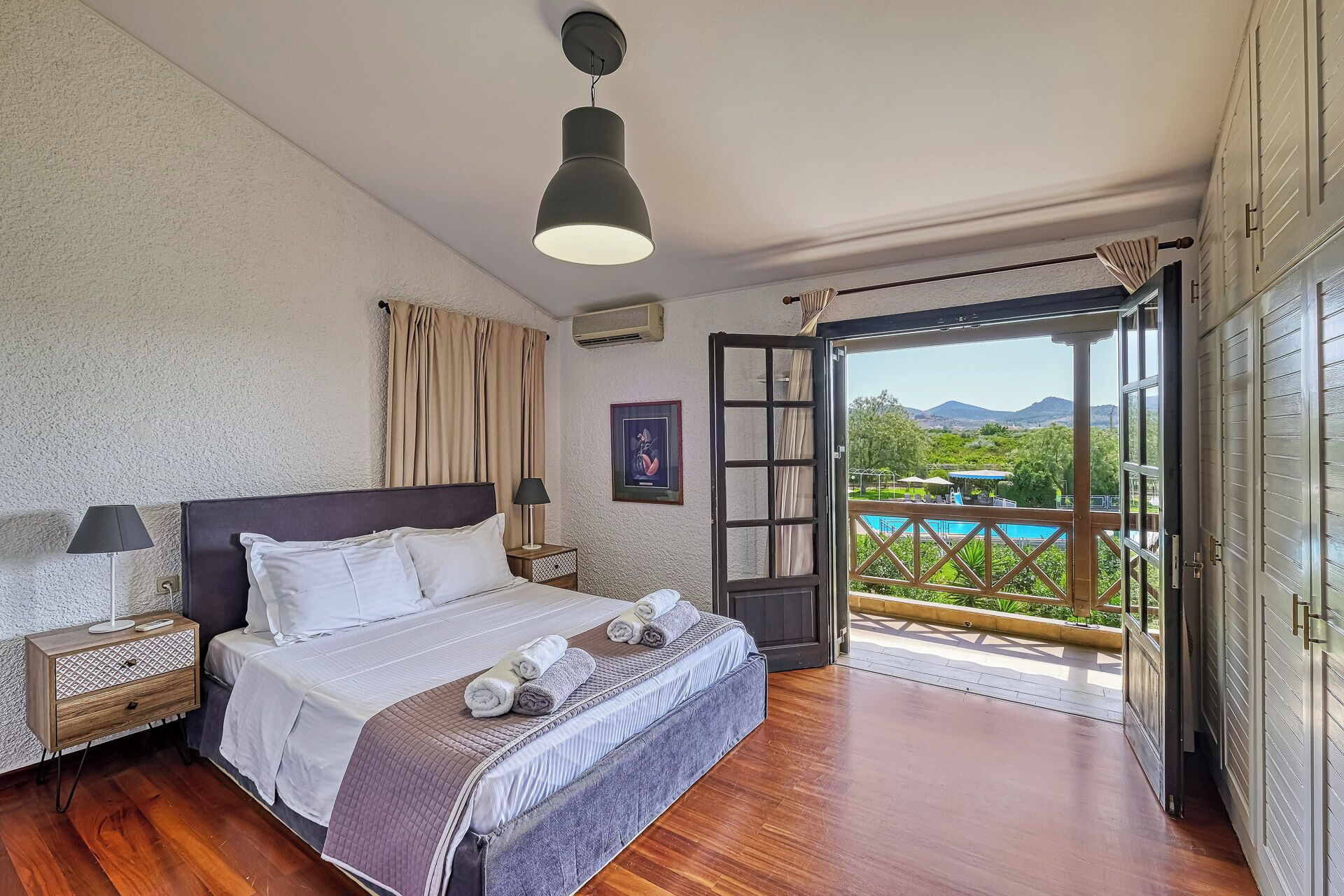 A bedroom with a bed, bedside tables, a nightstand, a window with curtains, and a view of a pool and mountains.