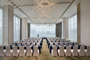Un salón de conferencias con filas de sillas blancas, mesas con manteles azules y un techo con luces colgantes.
