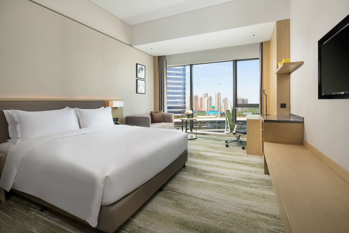 A hotel room with a large bed, a desk, a chair, a TV, and a view of the cityscape.