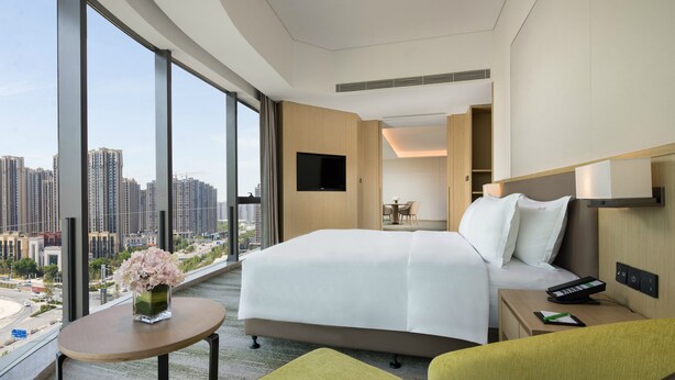 A modern hotel room with a large bed, a small table with a vase of flowers, a seating area with green chairs, a TV mounted on the wall, and a city view through large windows. - Holiday Inn Zhengzhou Riverside by IHG (Zhengzhou)
