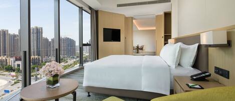 A modern hotel room with a large bed, a small table with a vase of flowers, a seating area with green chairs, a TV mounted on the wall, and a city view through large windows.