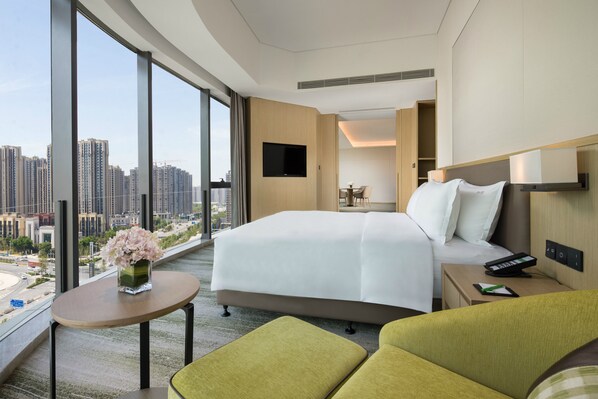 Habitación de hotel moderna con una cama grande, una mesita con un florero, una zona de estar con sillas verdes, un televisor montado en la pared y vistas a la ciudad a través de amplios ventanales.