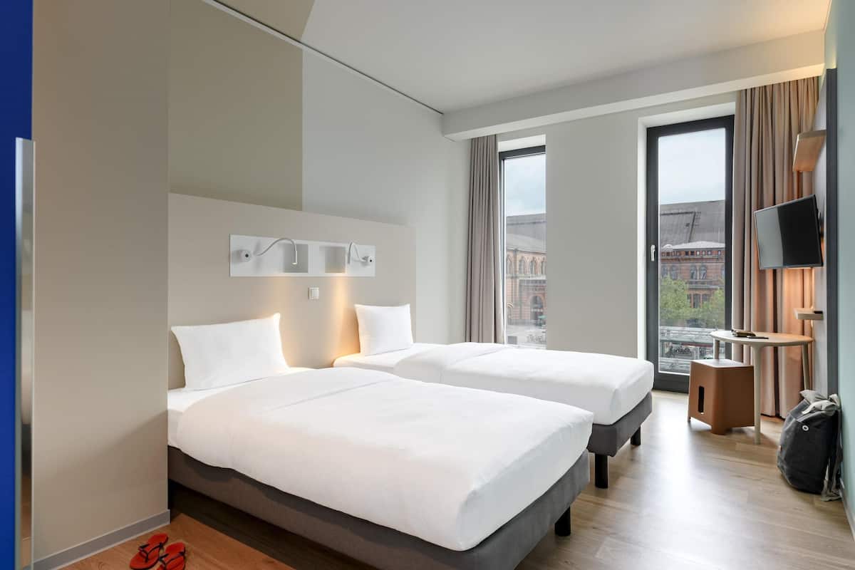 Habitación de hotel con dos camas, televisión, una mesita y vista a la ciudad.