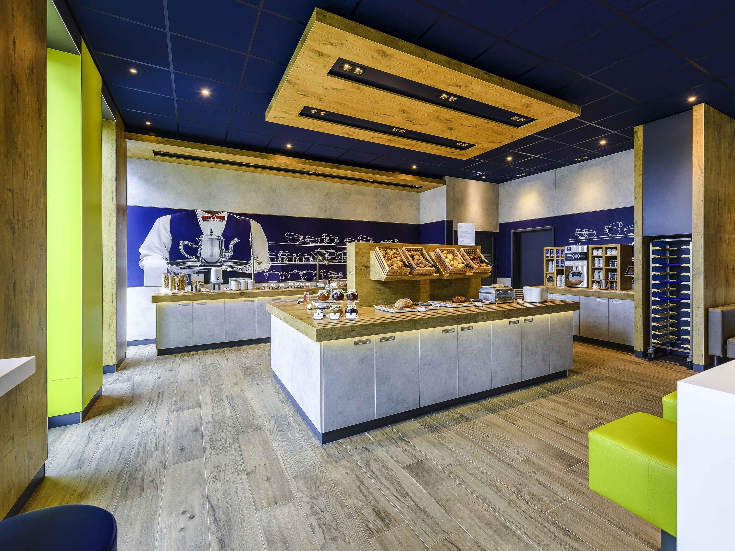 Un café moderno con una isla central que exhibe pasteles y café, una paleta de colores azul y amarillo, y un gran mural en la pared de fondo.