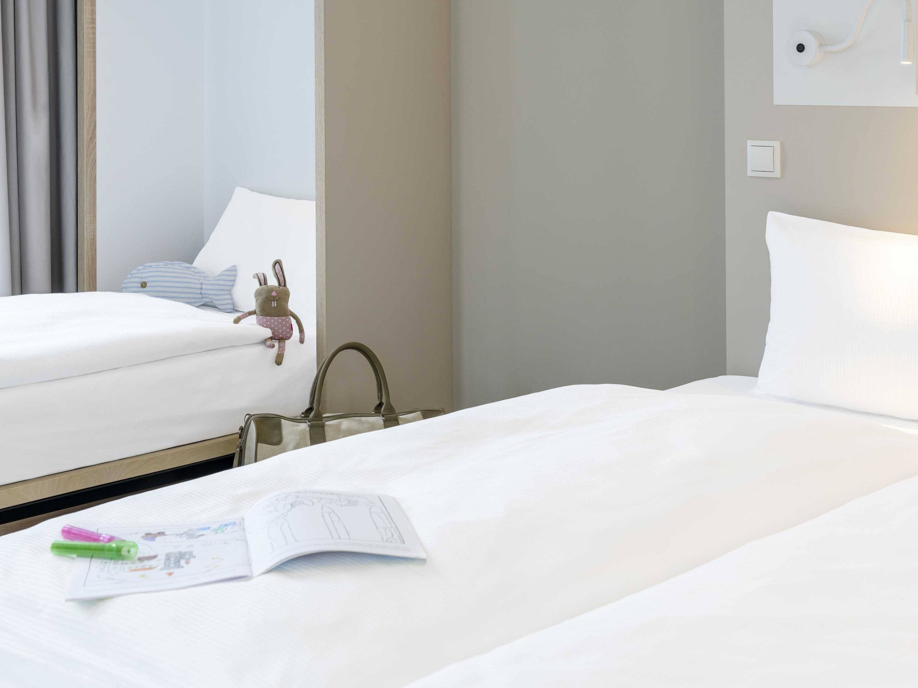 A hotel room with two beds, a plush toy, a handbag, and a wall-mounted light fixture.