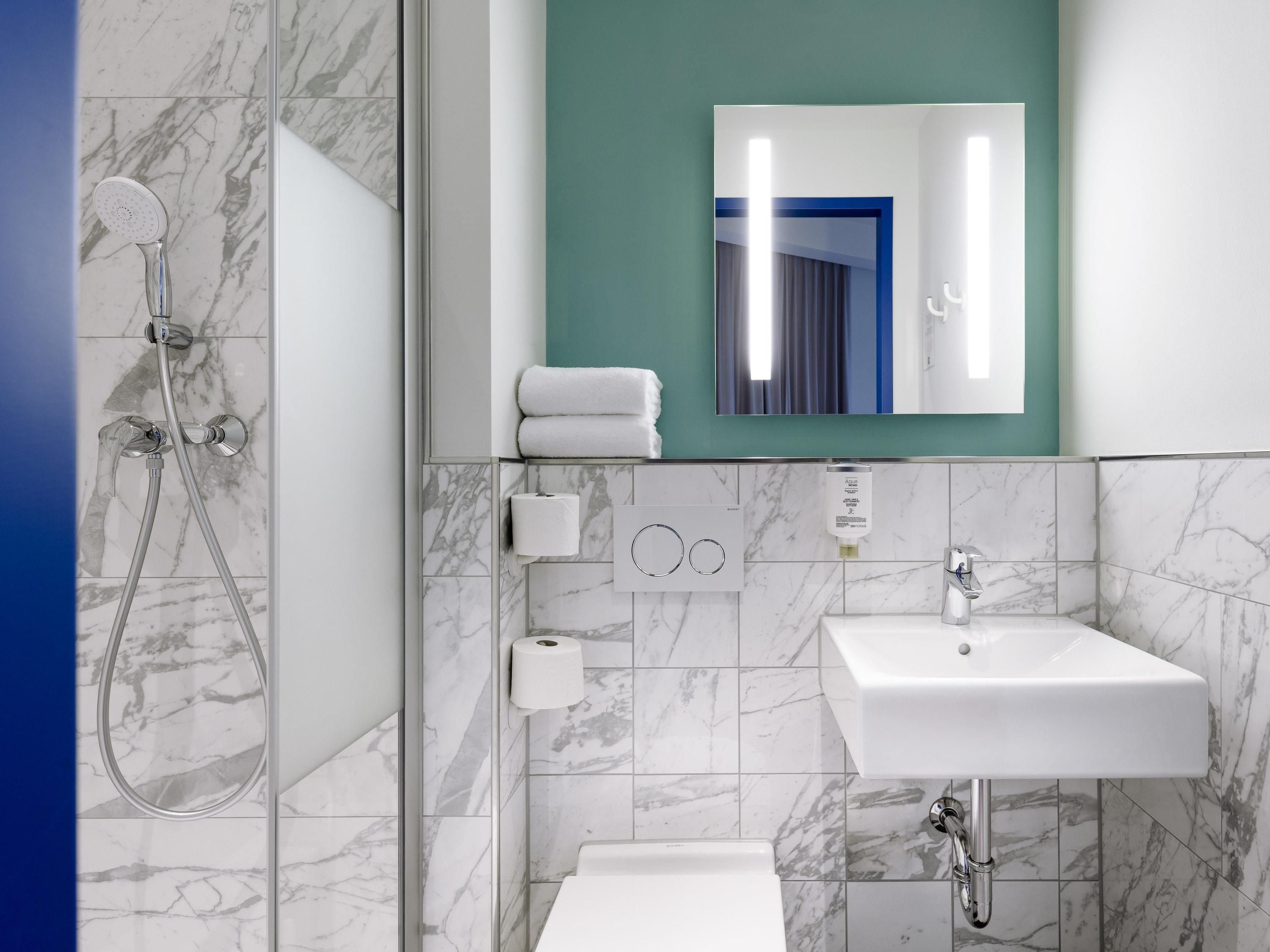 A modern bathroom with a shower, toilet, and sink, marble tiles, and a mirror with a view of a room.