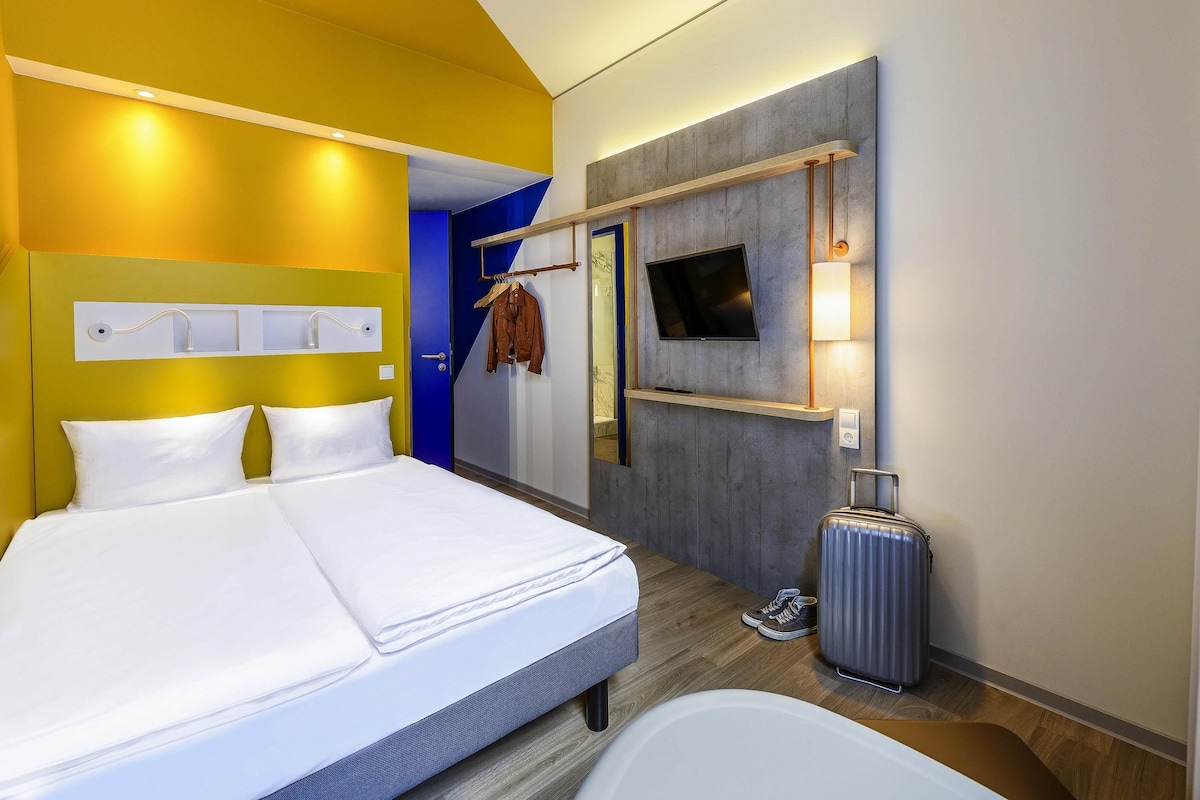 Una habitación de hotel con una cama, televisor, espejo y maleta.