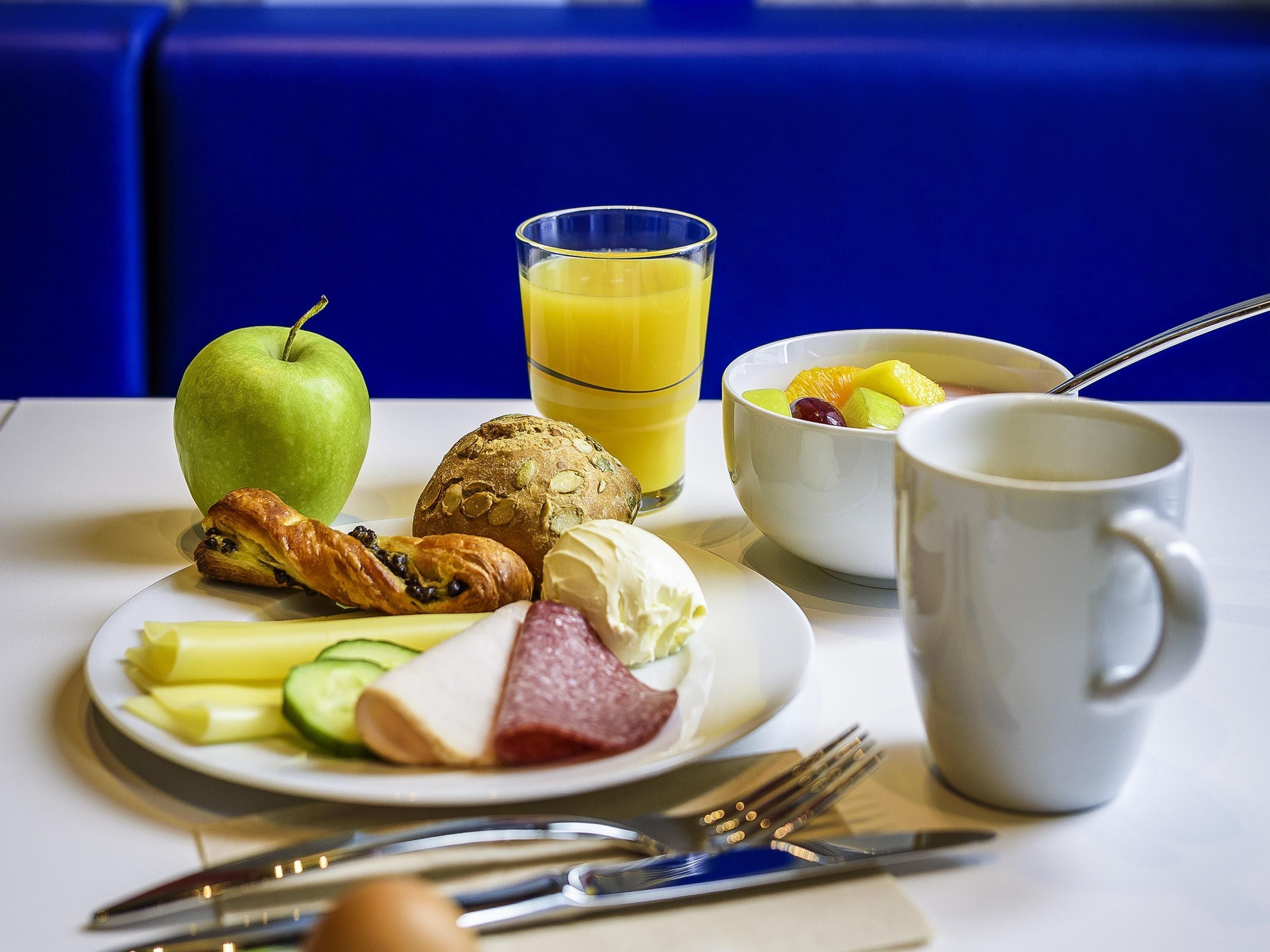 Un desayuno con un plato de comida, un vaso de jugo de naranja, un bol de fruta, un croissant, una loncha de jamón y una taza.