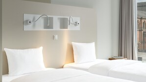 Une chambre d’hôtel avec deux lits, un luminaire mural moderne et une vue sur un immeuble par la fenêtre.