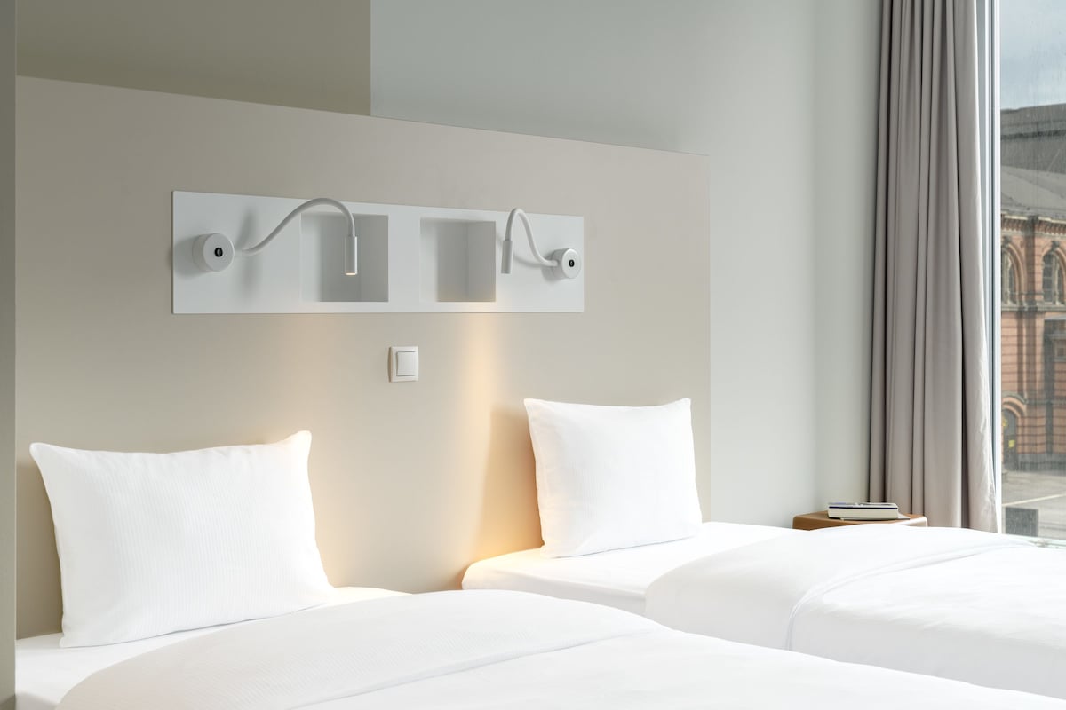 Een hotelkamer met twee bedden, een moderne wandlamp en uitzicht op een gebouw.