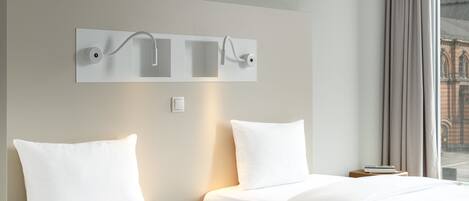 Habitación de hotel con dos camas, una lámpara moderna montada en la pared y vista a un edificio a través de la ventana.