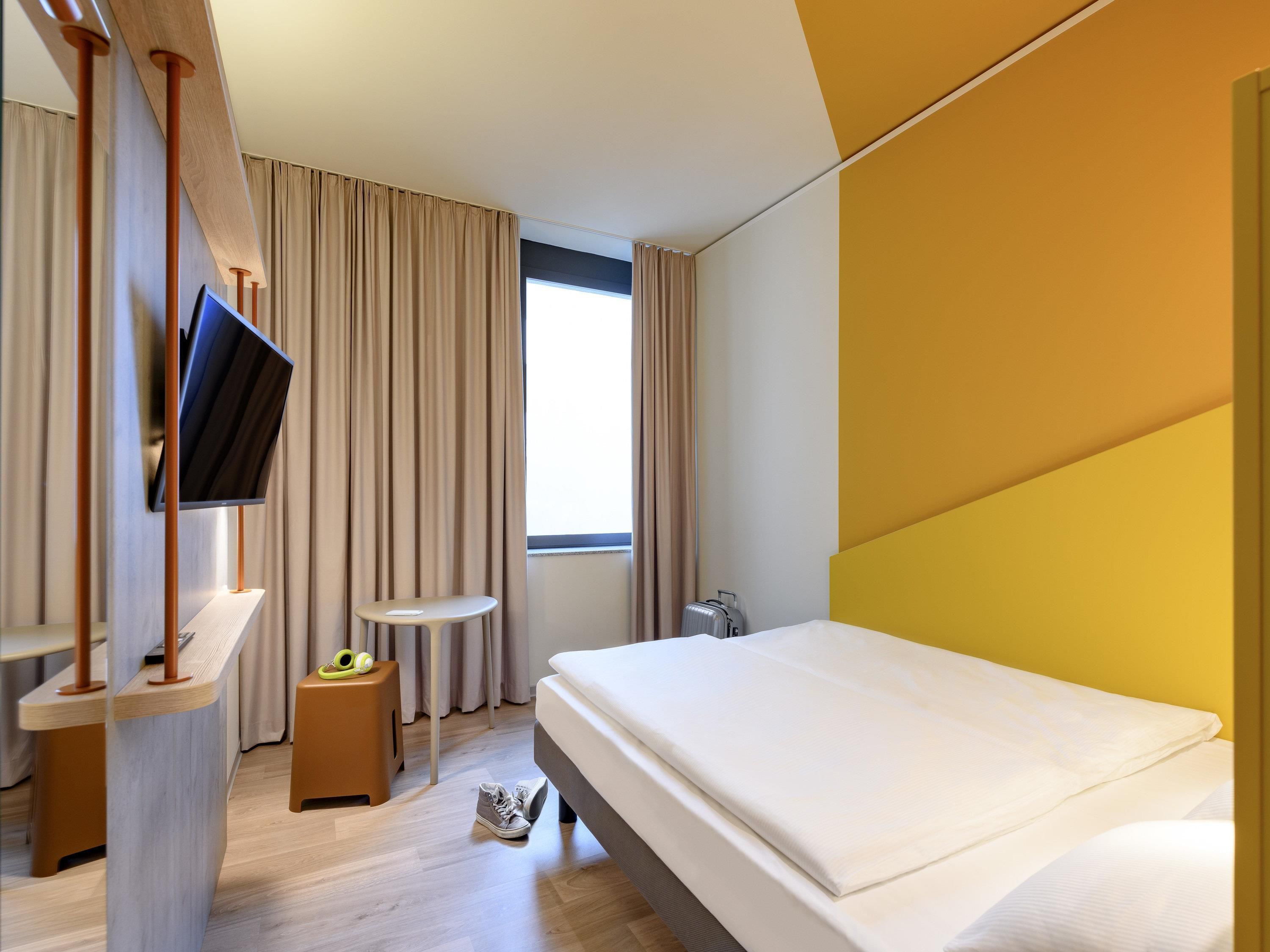 Habitació d'hotel moderna amb un llit, una televisió penjada a un panell de fusta, una taula rodona petita i una maleta.