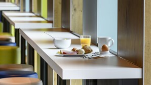 Preparación para el desayuno con un plato de comida, un vaso de zumo de naranja, una taza y un bol sobre una mesa blanca en una habitación moderna con detalles de madera y vistas a un pasillo.