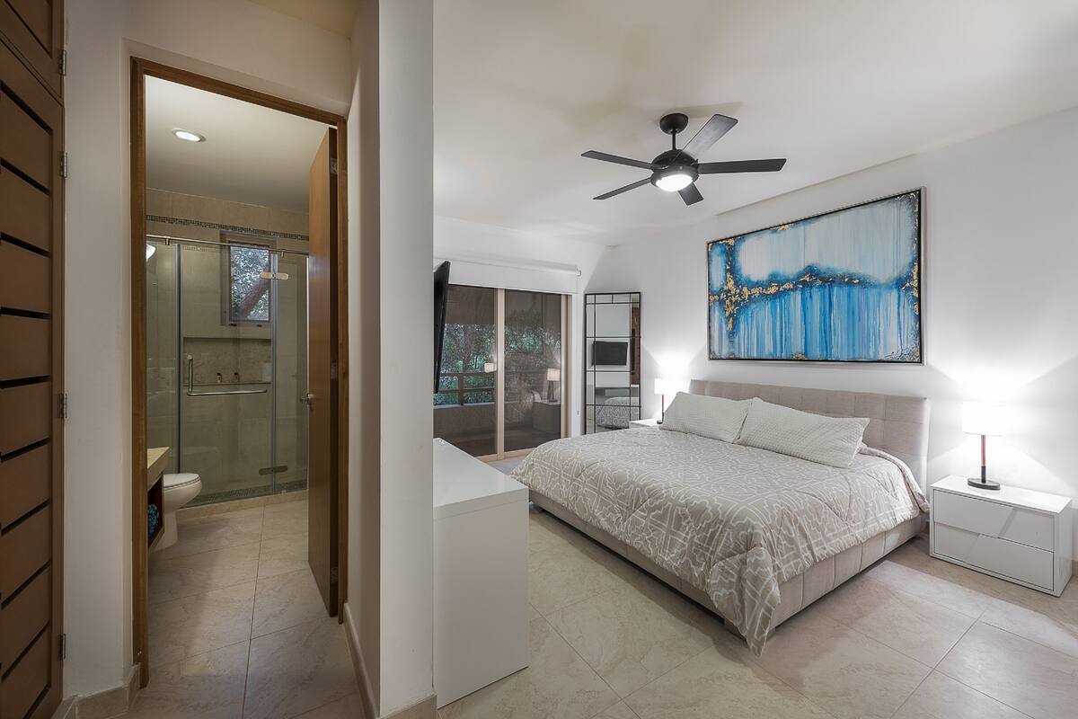 Un dormitorio con cama, ventilador de techo, mesita de noche y un cuadro en la pared.