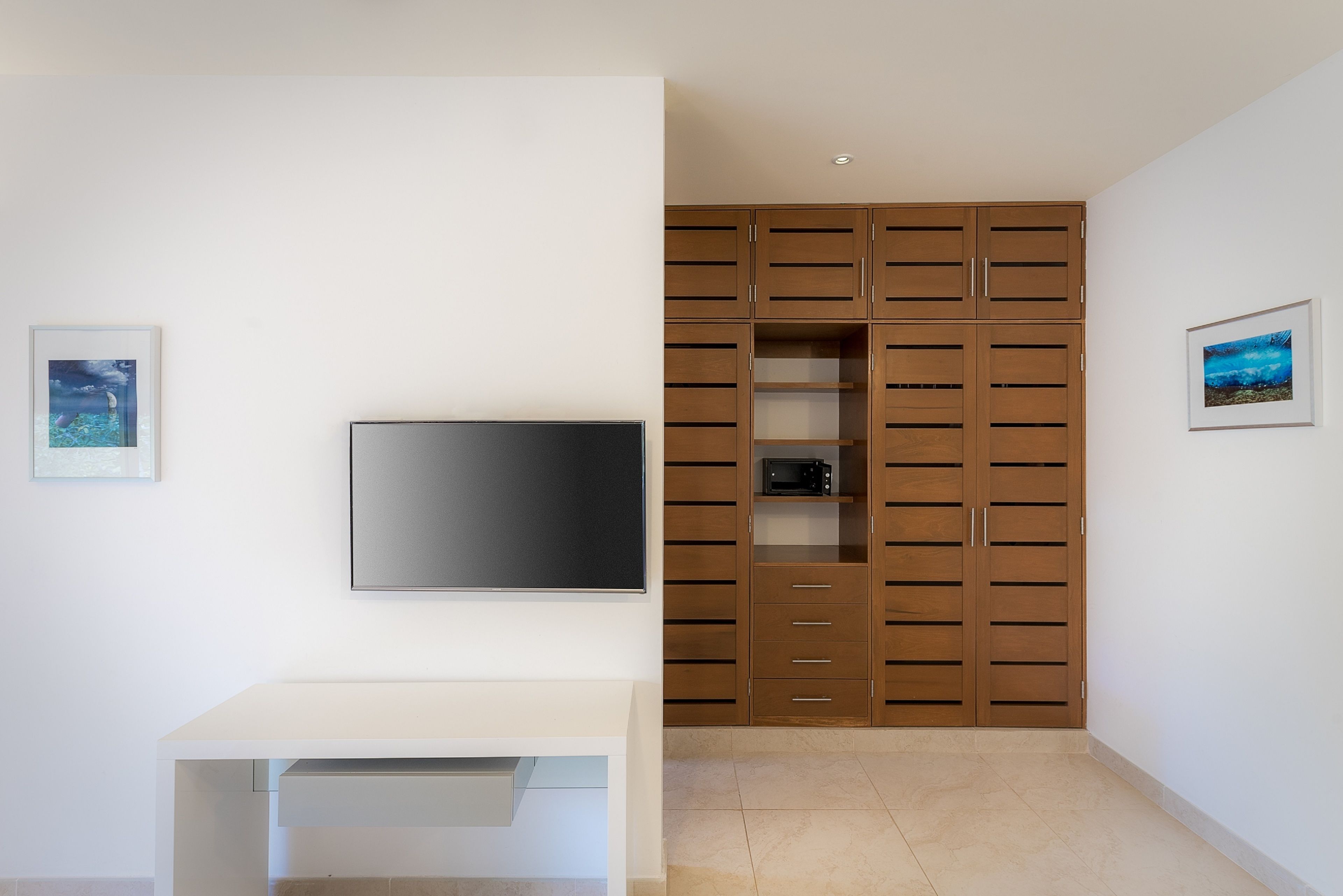 A modern room with a flat-screen TV mounted on the wall, a white desk, and a wooden cabinet with multiple drawers and shelves.