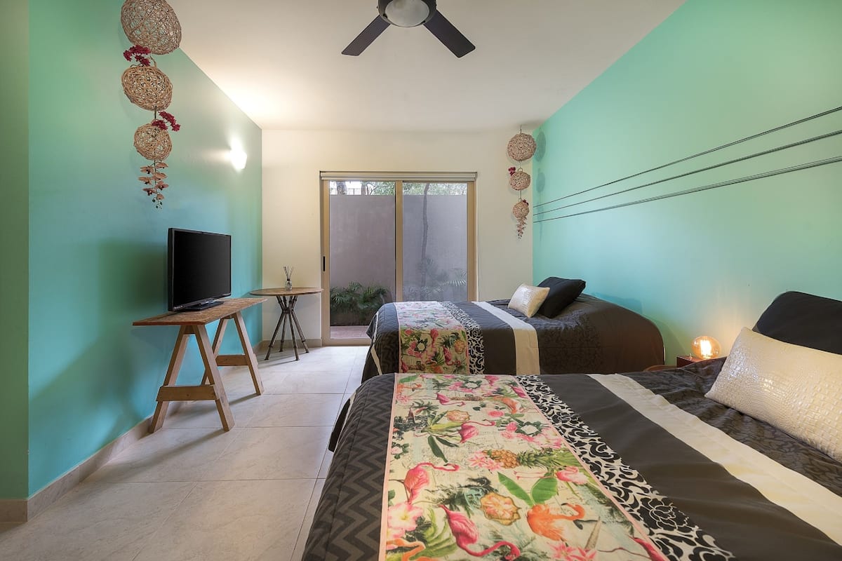 Un dormitorio con una cama, un televisor sobre un soporte de madera, un ventilador de techo y vistas al exterior por una puerta corrediza.