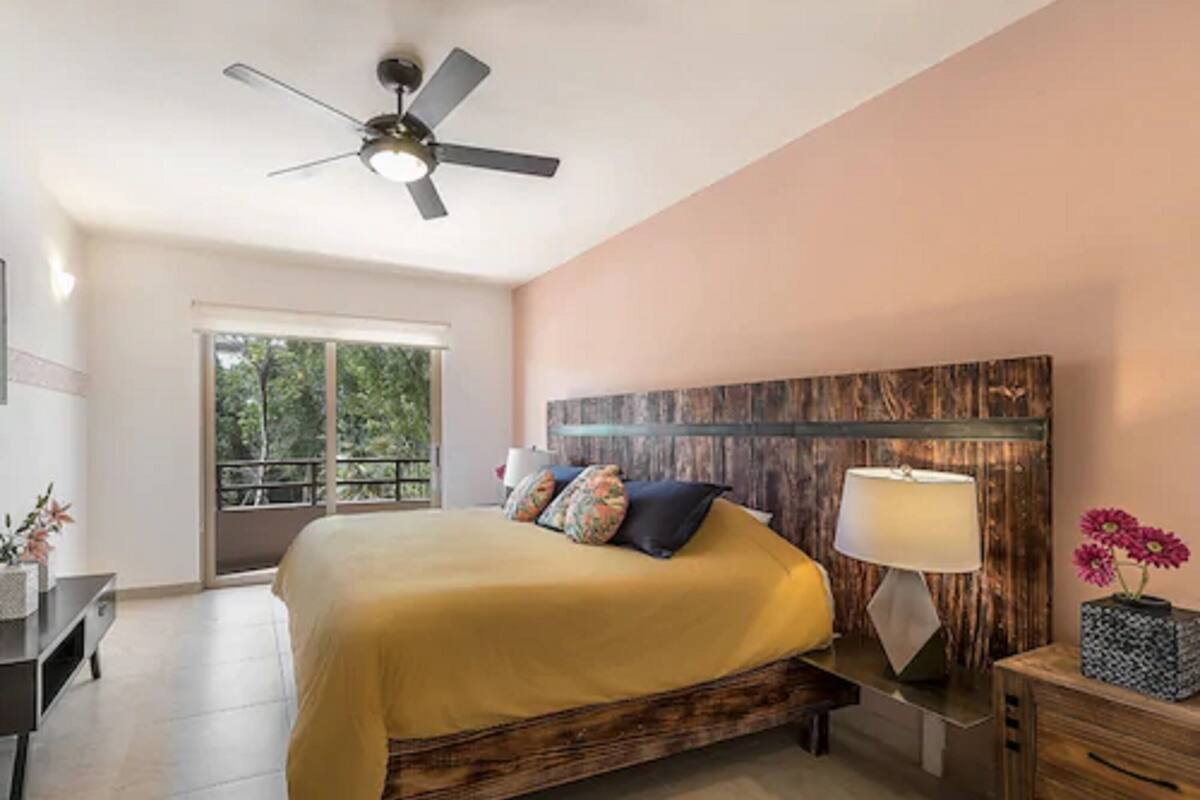 Un dormitorio con cama, mesita de noche, lámpara, cabecera de madera, ventilador de techo y balcón con vistas a árboles.