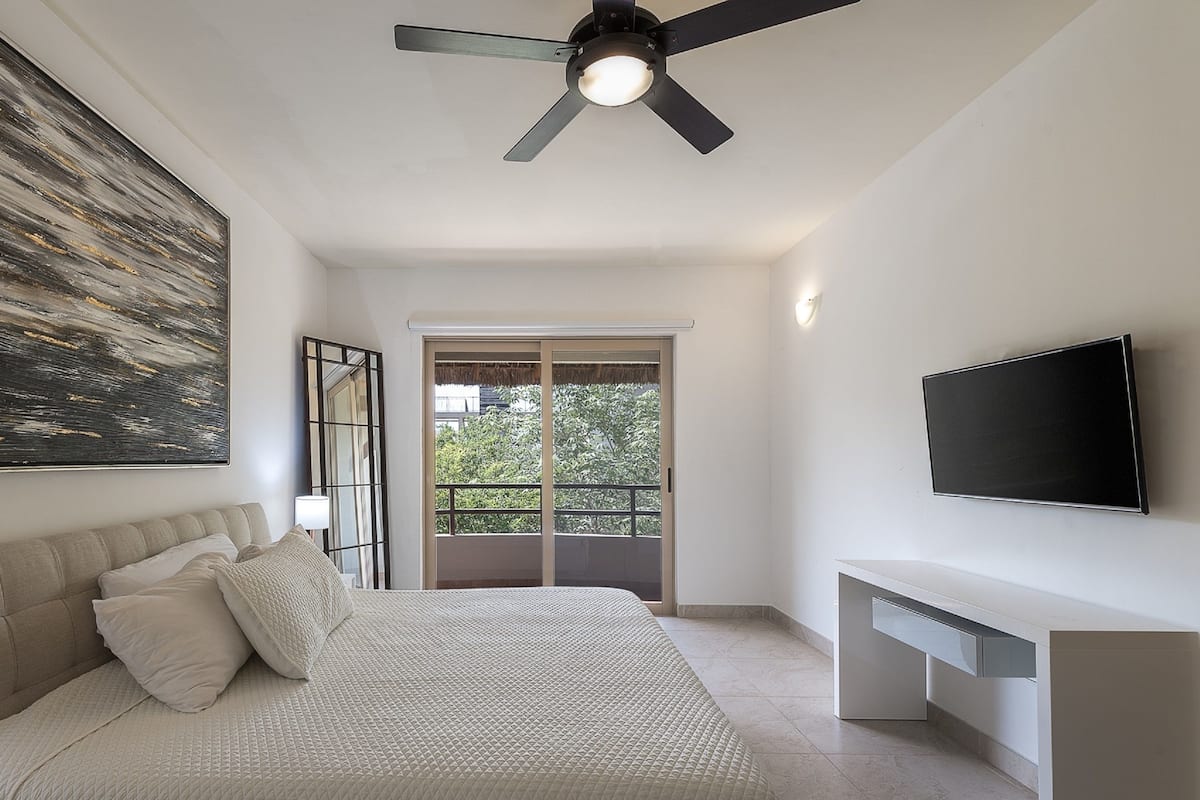 Un dormitorio moderno con una cama grande, ventilador de techo, televisor y balcón con vistas a zonas verdes.