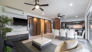 Una sala de estar moderna con un sofá, una mesa de centro y un televisor. Se ve una zona de comedor con sillas y una cocina al fondo.