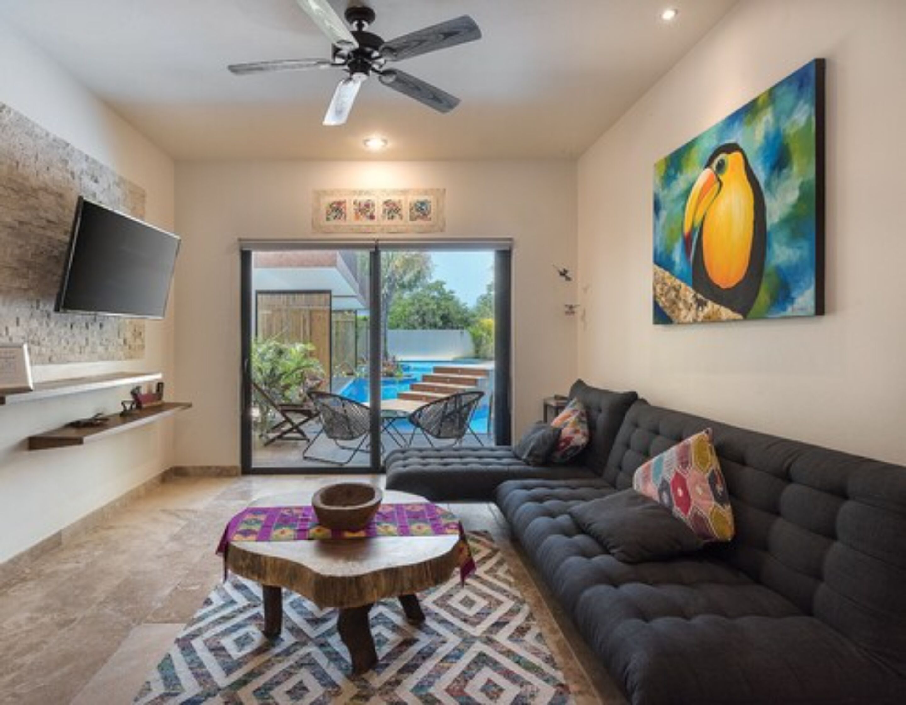 A living room with a dark sofa, a round wooden coffee table, a ceiling fan, a flat-screen TV mounted on the wall, and a painting of a toucan.