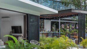 Un área de estar exterior moderna con barra, rodeada de abundante follaje y barandilla de vidrio.