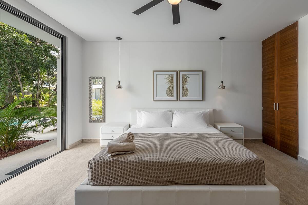 A modern bedroom with a large bed, two bedside tables, a wooden wardrobe, and a view of a garden through a glass door.