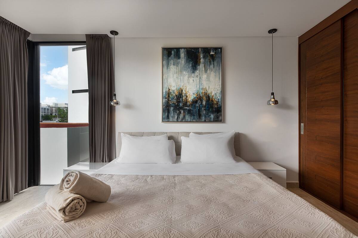 A modern hotel room with a large bed, a painting on the wall, and a view of the city through the window.