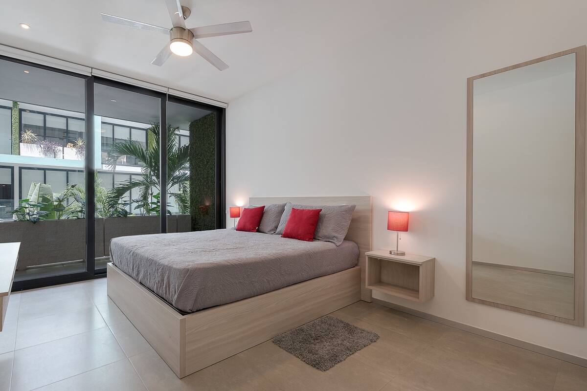A modern bedroom with a large bed, two bedside lamps, a ceiling fan, and a view of greenery outside.
