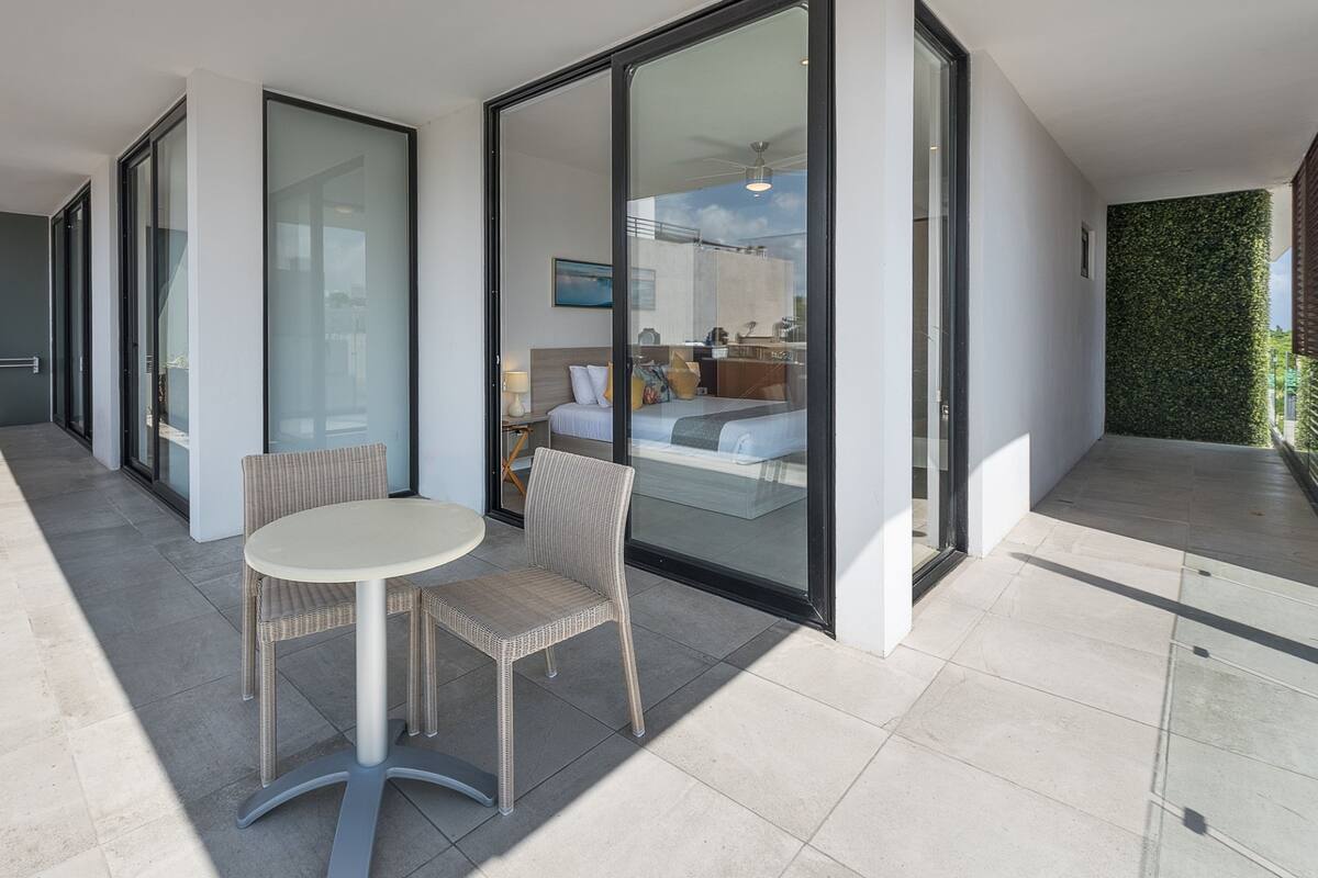 A modern hotel room with a glass door leading to a balcony, a round table, wicker chairs, and a bed with a view of the sky.